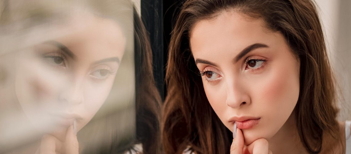 Adult woman in thoughtful pose looking out an urban window in natural light, evoking the introspection that accompanies an autism diagnosis received in adulthood